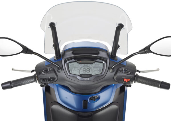 Close-up view of a scooter's dashboard, featuring a digital display showing speed and other metrics, with handlebars that include mirrors and various control switches. The scooter has a blue body and a transparent windshield.