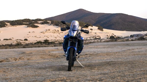 A blue motorcycle stands on a sandy terrain, facing the camera, with hills and dunes in the background under a clear sky.