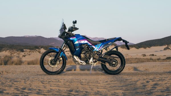A blue adventure motorcycle parked on sandy terrain, with mountains in the background and a clear sky.