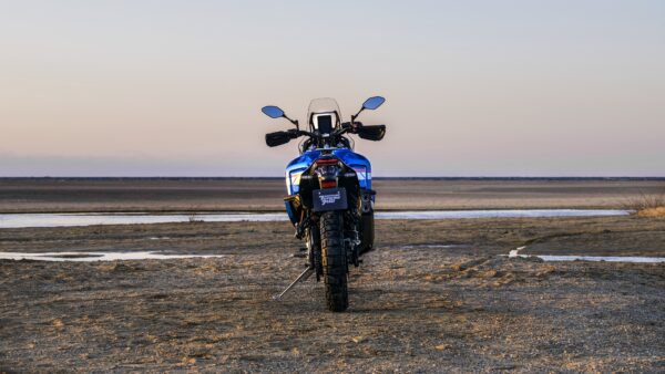 A blue motorcycle is parked on sandy terrain with a river in the background, under a soft sunset sky.