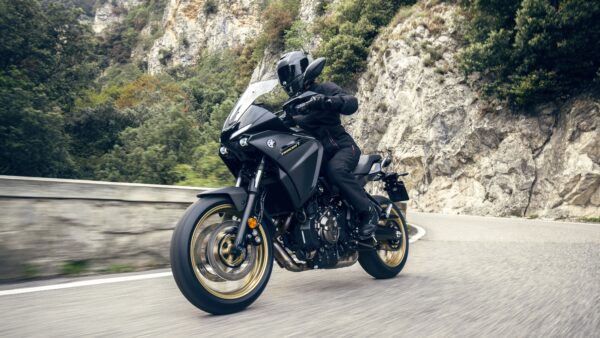 A rider in black gear is maneuvering a sleek, black motorcycle on a curvy road surrounded by greenery and rocky cliffs.