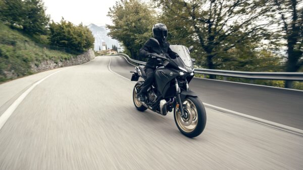 A motorcyclist in black gear rides a sleek motorcycle on a winding road, surrounded by trees and hills, with a sense of motion captured in the image.