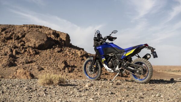 A blue and black adventure motorcycle stands on rocky terrain with a desert landscape in the background.