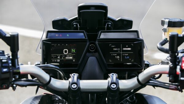 Close-up view of a motorcycle's dashboard displaying digital gauges, including speed (0 km/h), trip distance (390.3 km), temperature readings, and various warning indicators, with handlebars and controls visible.