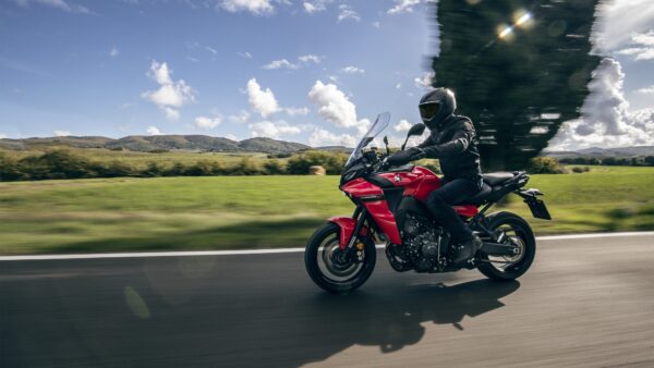 A motorcyclist in a black helmet and jacket rides a vibrant red motorcycle on a scenic road, with lush green fields and mountains in the background under a partly cloudy sky.