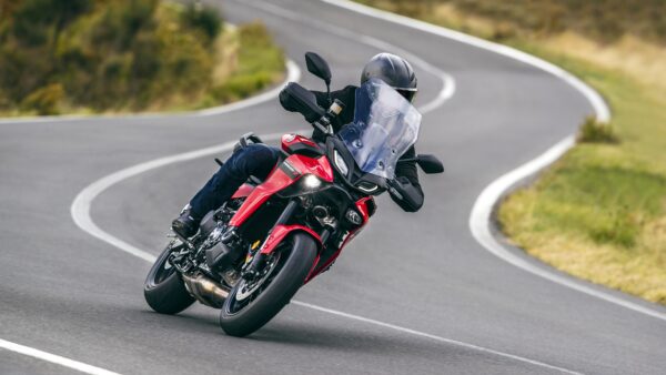 A rider in black gear leans into a curve on a red motorcycle with a large clear windscreen on a winding road surrounded by greenery.
