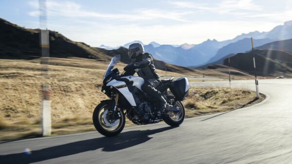 A motorcyclist in protective gear rides a white Yamaha motorcycle on a winding road with mountains in the background.