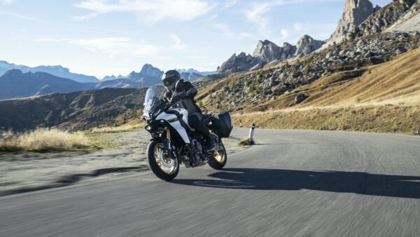 A motorcyclist in a black helmet and gear rides a sleek, white motorcycle along a winding road in a mountainous landscape, with rocky peaks and rolling hills in the background under a clear blue sky.