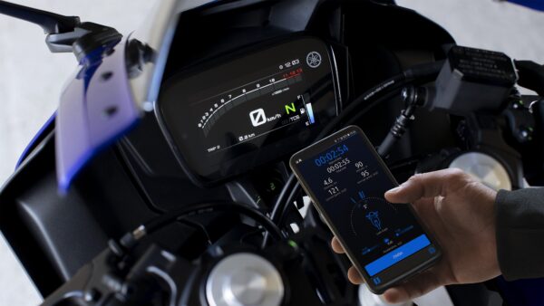 A close-up view of a motorcycle dashboard displaying speed and trip information, alongside a smartphone showing data such as time, lean angle, and speed metrics.