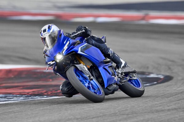 A racer in black gear leans into a turn on a bright blue sport motorcycle, with the track's curbing visible beneath them.
