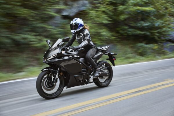 A rider in a black motorcycle suit and blue helmet accelerates on a sleek black sportbike along a winding road, surrounded by lush greenery.