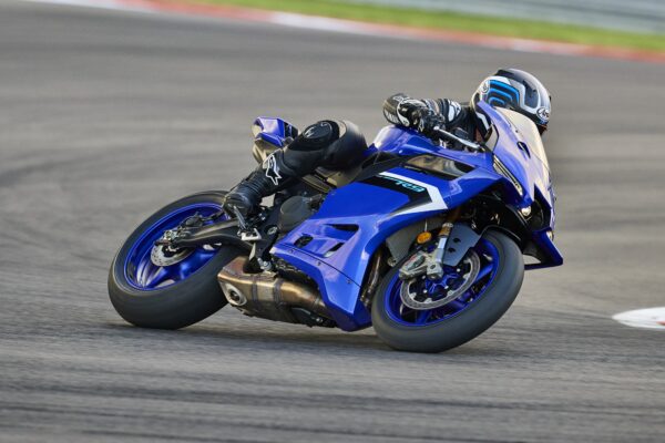 A motorcyclist leans into a turn on a blue sport motorcycle, wearing a black racing suit and helmet, on a racing track.