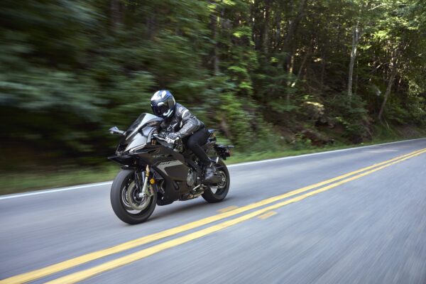 A rider in black gear rides a sleek black motorcycle on a winding road, surrounded by lush green trees, capturing a sense of speed and motion.