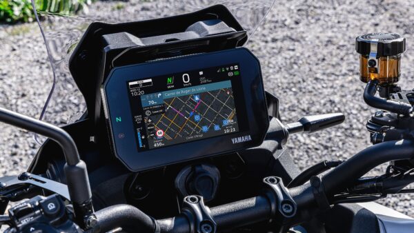 A close-up view of a Yamaha motorcycle's dashboard featuring a digital navigation display showing a map and navigation instructions, alongside control handles and a brake fluid reservoir. The surroundings are blurred, suggesting an outdoor setting.