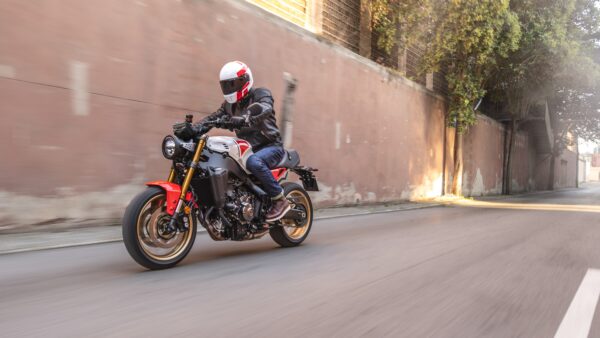 A motorcyclist in a leather jacket and white helmet rides a stylish motorcycle with red and black accents, speeding down a city street with a blurred background.