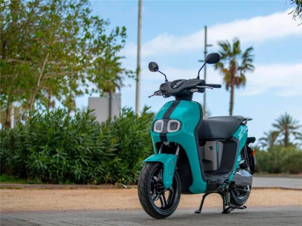 A turquoise electric scooter parked in a landscaped outdoor area with palm trees and a blue sky in the background.