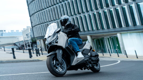 A rider in a black jacket and helmet is leaning into a turn on a modern scooter, set against a backdrop of contemporary glass buildings.