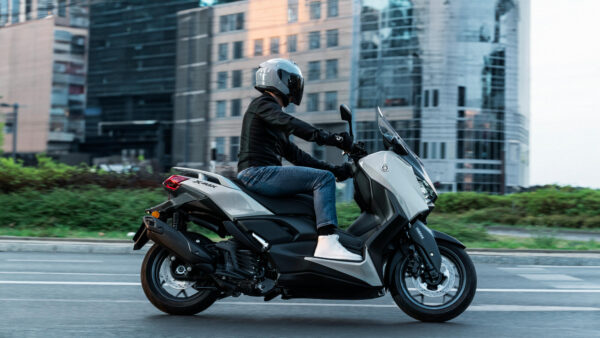 A rider wearing a gray helmet and black jacket is riding a gray Yamaha Xmax scooter on a city street with modern buildings in the background.