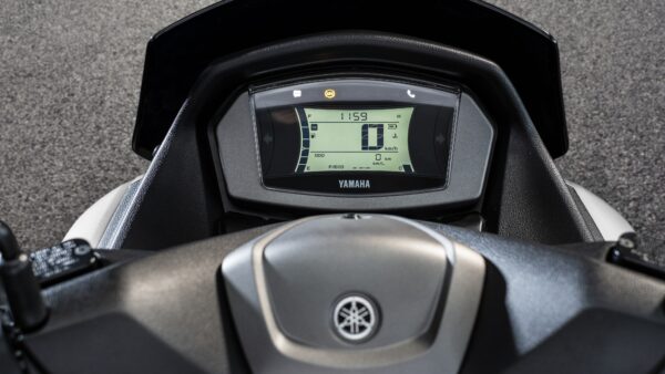 A close-up view of the dashboard of a Yamaha scooter, displaying an LCD screen with various indicators, including speed, fuel level, and odometer readings, against a textured gray background.