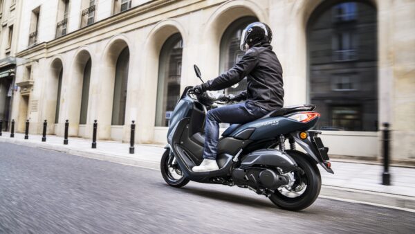 A person wearing a helmet and a black jacket rides a gray scooter on a city street, passing by elegant buildings and black bollards.