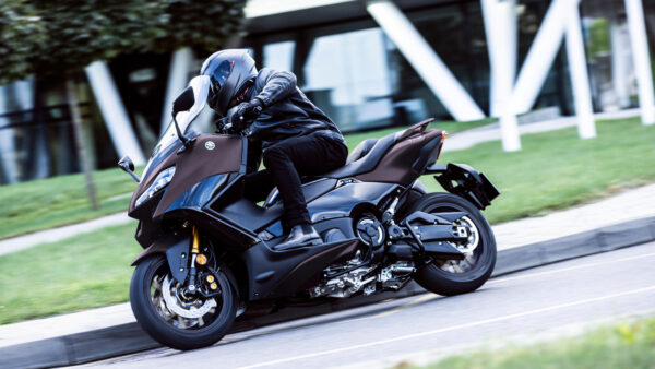 A motorcyclist in a black helmet and leather jacket is leaning forward while riding a sleek, dark motorcycle on a curved road, with modern architecture and greenery in the background.