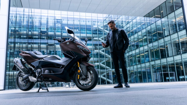 A man in a black leather jacket stands beside a brown Yamaha motorcycle, looking at his phone, with a modern glass building in the background.