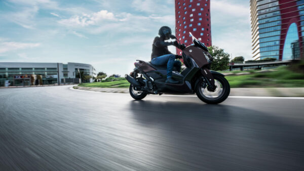 A motorcyclist in a black helmet rides a dark scooter around a curve on a smooth road, with modern buildings and a blue sky in the background.