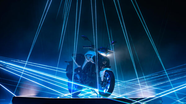 A white motorcycle is illuminated by blue laser beams in a dark environment, creating a dramatic effect.