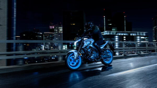 A motorcyclist in black gear rides a sleek, modern motorcycle with glowing blue wheels on a bridge at night, with a city skyline illuminated in the background.