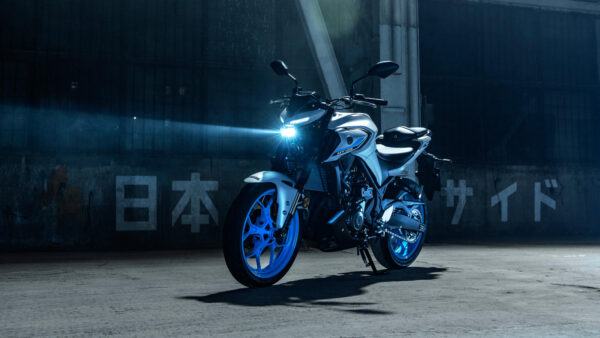 A modern motorcycle with a sleek design is illuminated by a bright headlight, set against a dark, industrial backdrop featuring Japanese characters. The motorcycle has a white body with blue accents on the wheels.