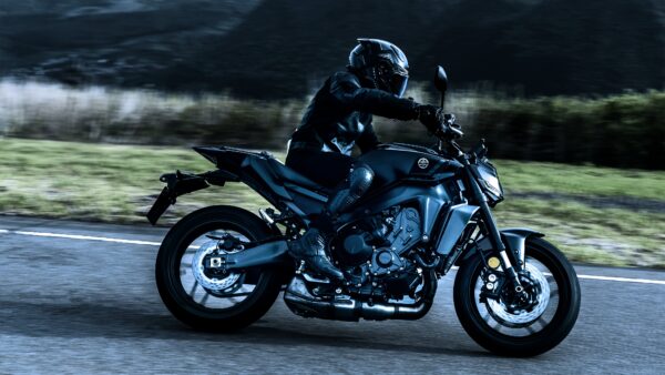 A motorcyclist in black gear rides a sleek blue motorcycle on a winding road, surrounded by lush greenery and distant hills.