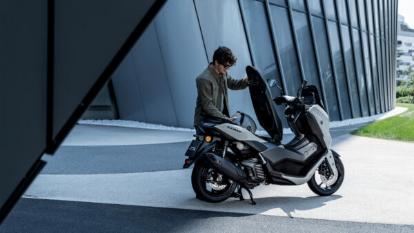 A man dressed in a green jacket is interacting with a sleek gray scooter in a modern architectural setting, with a curved building in the background and manicured greenery nearby.