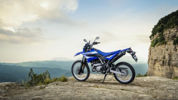 A blue dirt bike parked on a rocky outcrop with mountains and a cloudy sky in the background.