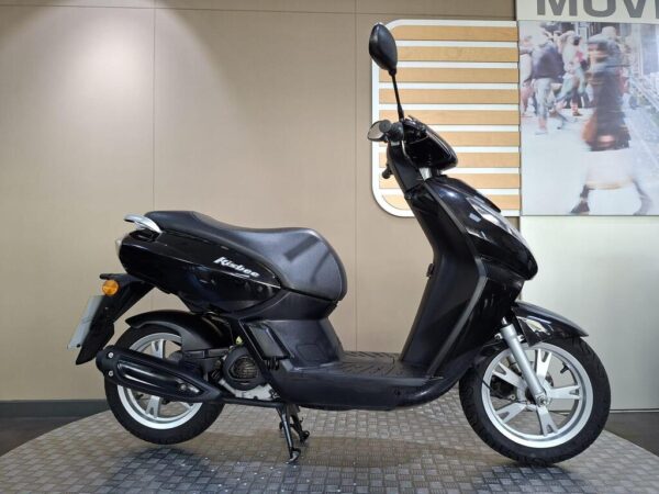 Peugeot Kisbee 50 (2018) A black scooter is parked on a textured surface, showing its side profile with a sleek design. The background features a blurred image of people walking.