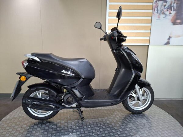 A black scooter with a sleek design is displayed on a textured surface, showcasing its profile and handlebars. The background is a neutral-colored wall with minimal decor.