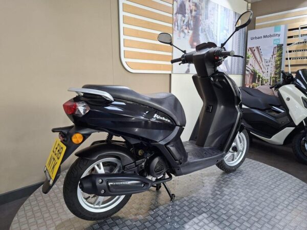 A black scooter is displayed in a showroom, featuring a sleek design with a comfortable seat, visible wheels, and a license plate. The background includes promotional materials related to urban mobility.