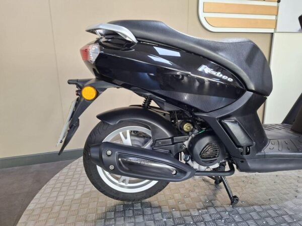A black scooter is parked on a textured surface, showing its rear side, seat, and exhaust. The model name "K-Slee" is visible on the body, with a small orange signal light near the rear.