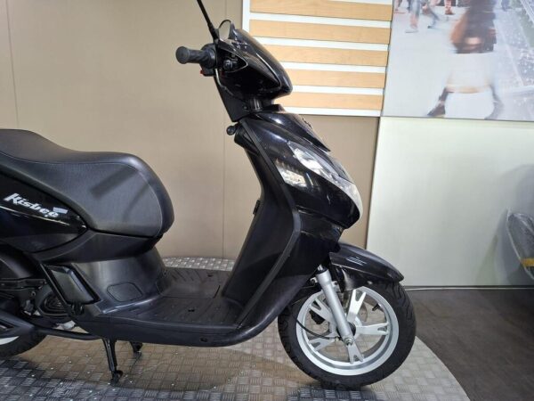A black scooter is displayed in profile view, featuring a sleek design, a comfortable seat, and front wheels. The background has a light-colored wall and a blurred image of people walking.