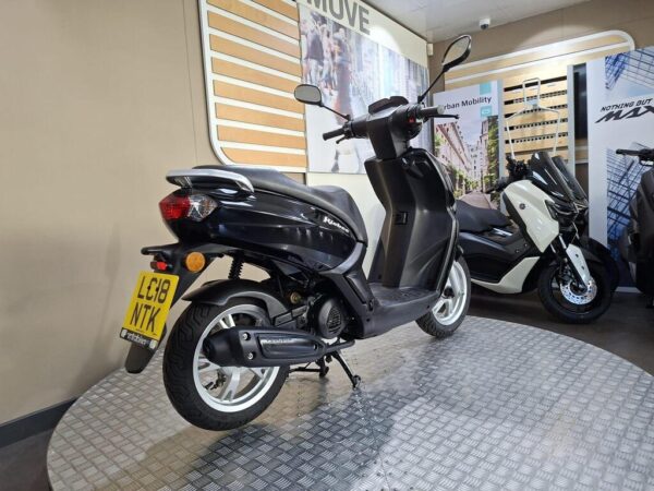 A black scooter with a visible license plate reading "LGB NTK" is displayed in a showroom, with a textured circular platform beneath it and additional scooters in the background.