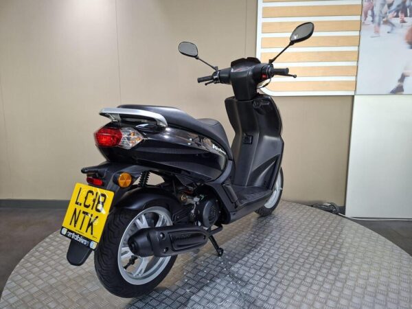 A black scooter displayed on a circular metallic platform, showing its rear and side profile with a visible license plate.