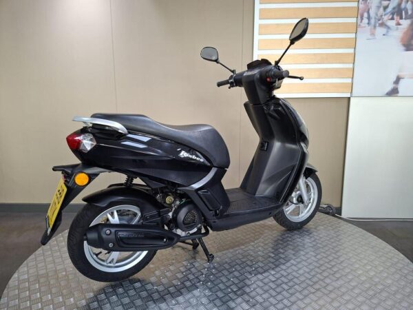 A black scooter parked on a textured platform, showcasing the side view with a focus on its sleek design and features. The background displays a neutral-toned wall with a glimpse of a patterned surface.