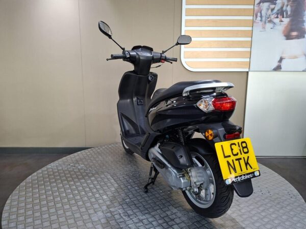 A black scooter positioned on a circular display platform, showing its rear and side view, with a license plate visible. The background features a muted wall with a decorative panel.
