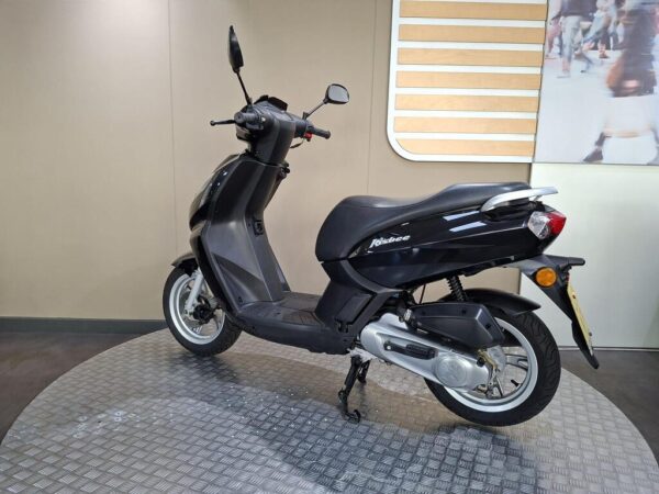 A black scooter is displayed on a circular platform, featuring a sleek design with a low seat and large wheels. The background is a neutral tone with wooden accents.