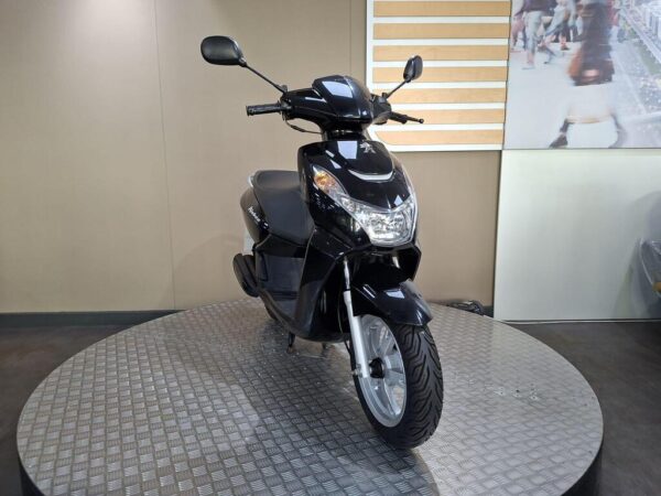 A black scooter displayed on a circular metal platform in a well-lit showroom, with beige walls and a background image of pedestrians.