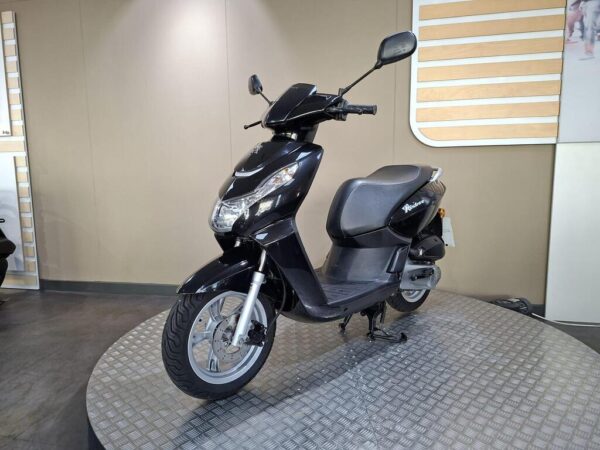 A black scooter displayed on a circular metallic platform in a showroom, with a neutral backdrop and wooden accents.
