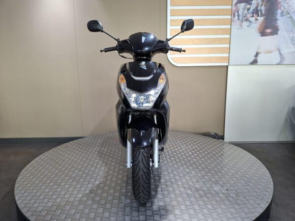 A black scooter positioned on a circular metallic platform, facing forward. The background features a neutral wall and part of an image showing people.