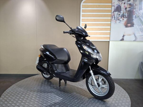 A black scooter is positioned on a circular, textured platform against a neutral background, with a blurred image of people in motion on the wall behind it.