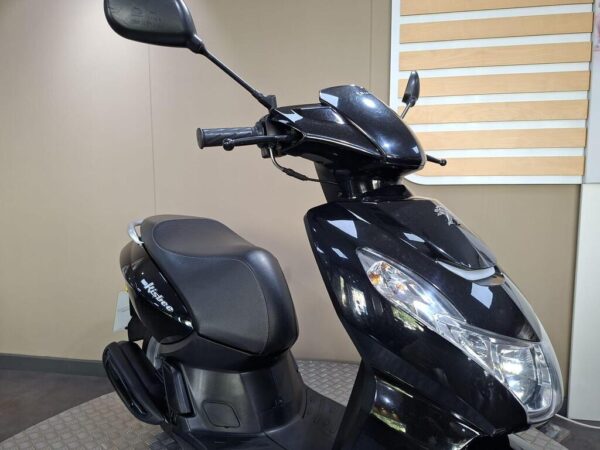 A black scooter displayed in a showroom with a sleek design, featuring a comfortable seat, handlebars, and rearview mirrors. The background is neutral-colored, emphasizing the scooter's features.