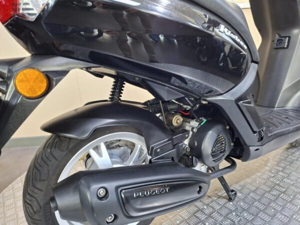 Close-up view of the rear section of a black scooter, highlighting the rear wheel, exhaust system, and engine components. The image shows a textured metallic surface beneath the scooter.
