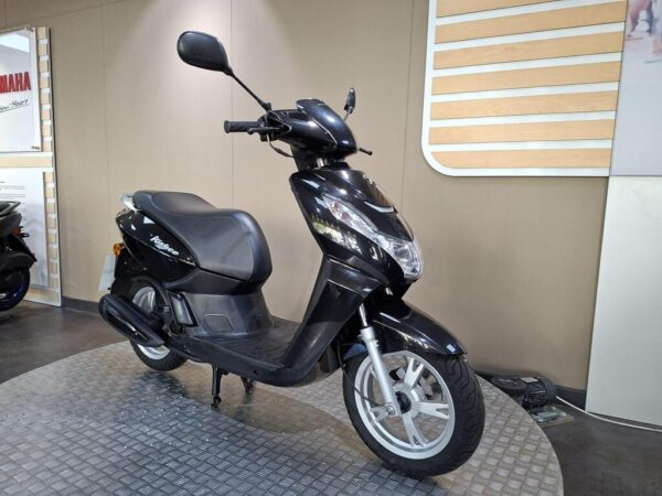A black scooter displayed in a showroom with a textured circular platform, featuring a sleek design and visible brand logos in the background.